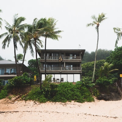 a photo taken from on a beach facing a large, 3 level home, with stairs leading up to it, surrounded by trees and bushes