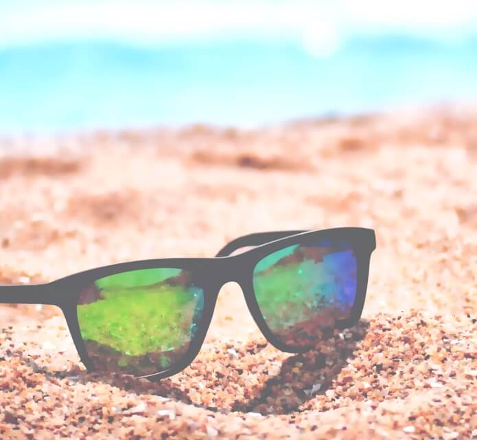 a pair of sunglasses, sitting in the sand on a very sunny day with a blue background that could be the water