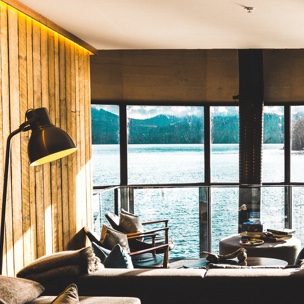 a photo of the inside of a cabin over looking the water, with a lamp a couch and chairs all visible in the photo